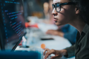 Focused black female programmer wearing glasses while coding new computer language while working on desktop PC in the office.