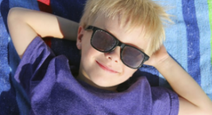 Young blonde boy laying on a towel outside with protective sunglasses on.