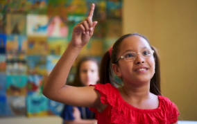Young girl with glasses raising her hand in class.